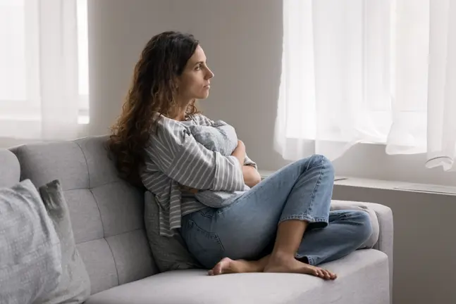 Pensive young woman seated on a couch at home, reflecting deeply while hugging a cushion and gazing out a window. This image symbolizes introspection and the process of contemplating the Judgment and Image in a step-by-step I Ching guide.